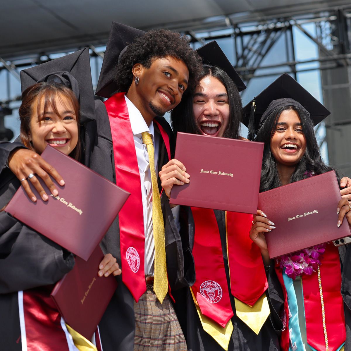Four smiling Santa Clara University graduates in caps and gowns celebrate with diplomas at commencement.