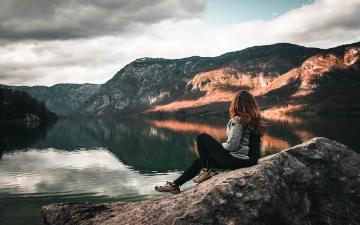 A woman sitting on a rock overlooking a still lake reflecting nearby mountain range