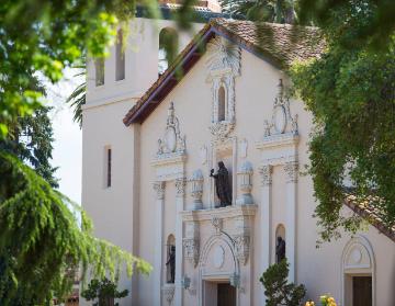 The image shows the front view of Mission Santa Clara surrounded by greenery.