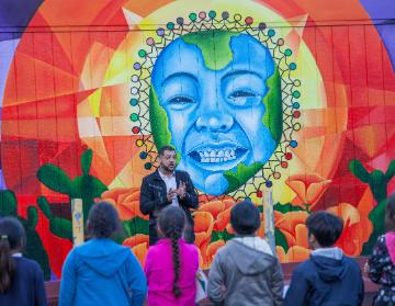 A person speaking to a small crowd in front of a colorful mural.