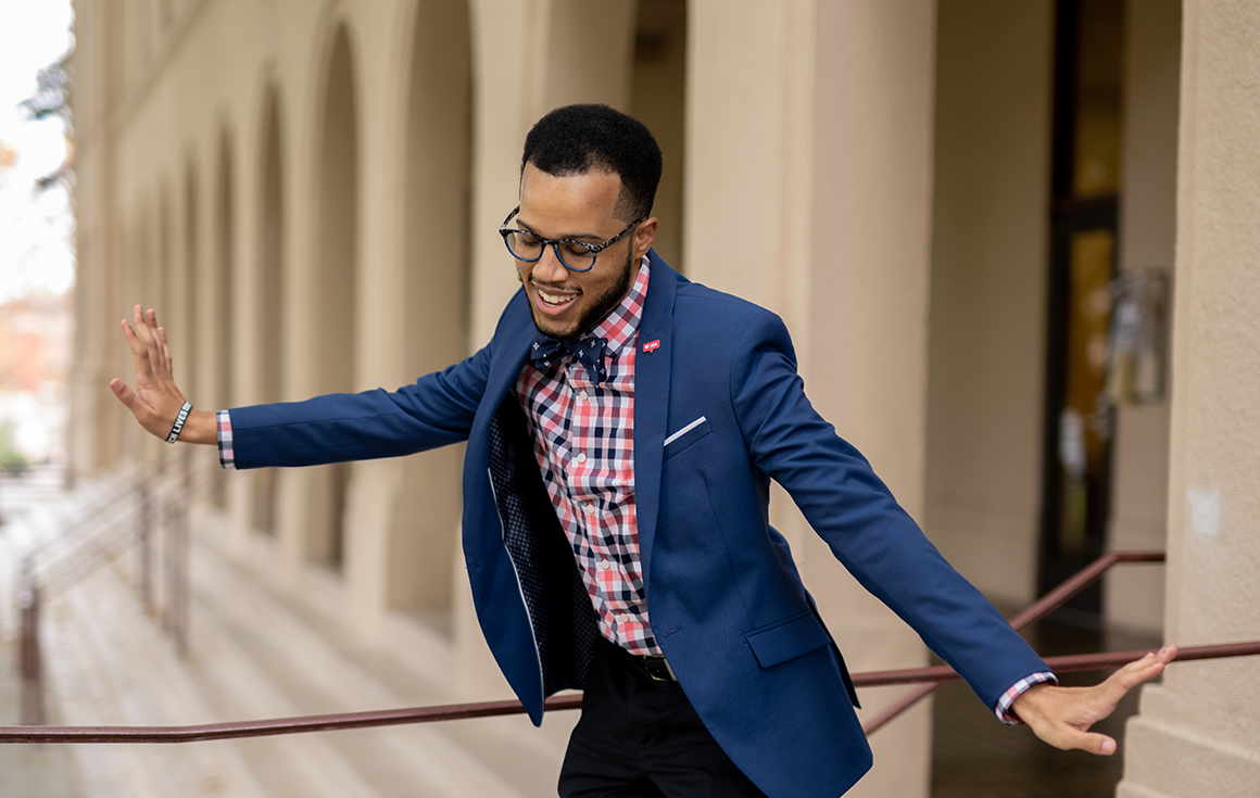 A Black man wearing a suit jacket, bowtie, and glasses stands, with arms playfully outstretched on the exterior steps to a building.