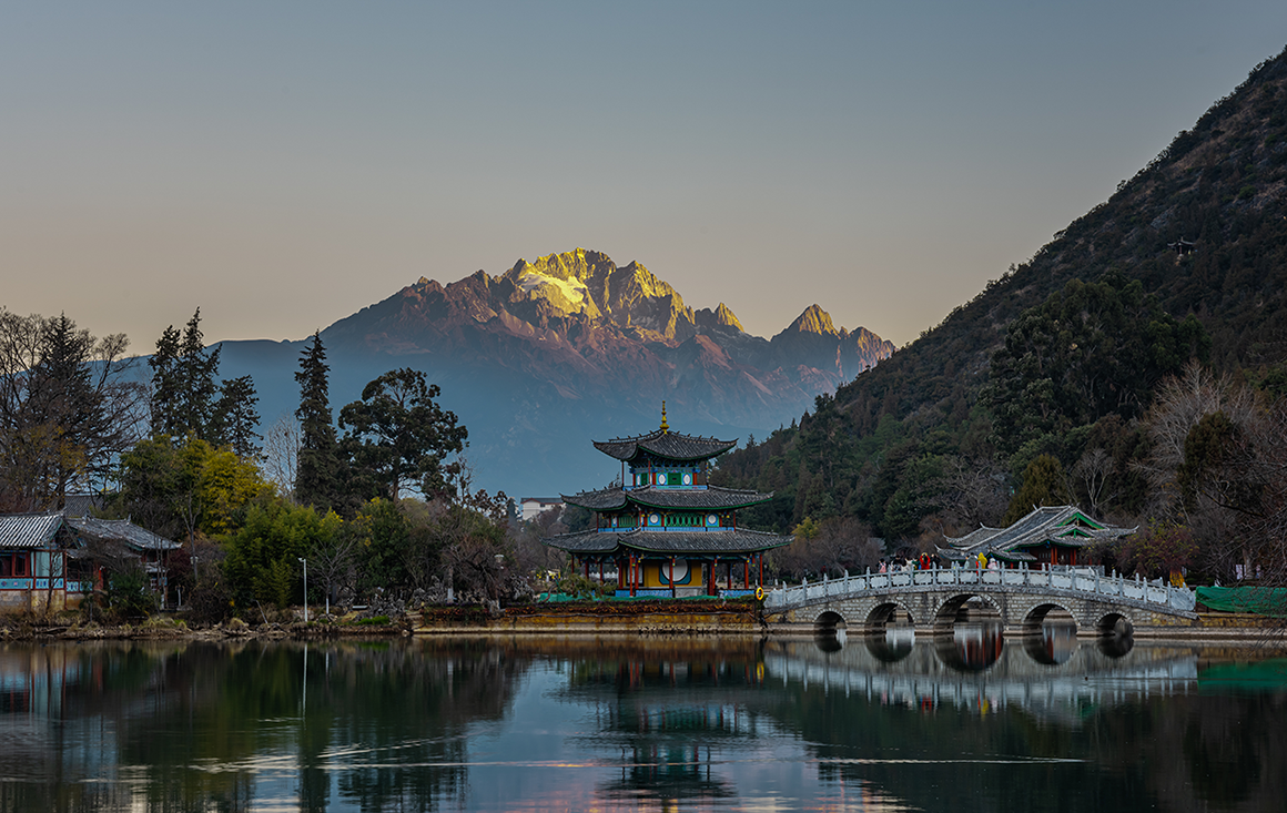 A peaceful pre-dawn morning looking over a still lake, a traditional Chinese pagoda, and majestic snow-capped mountains in the background.