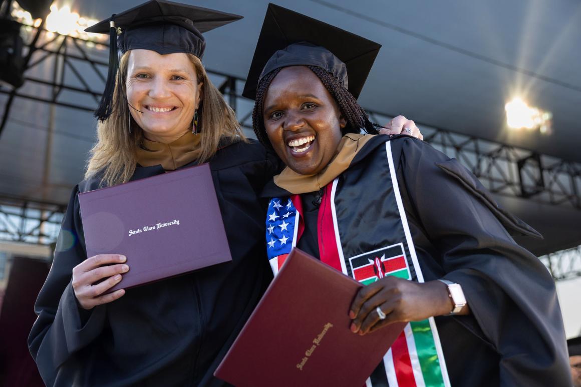 Two grad students smile while holding up their diplomas on stage.