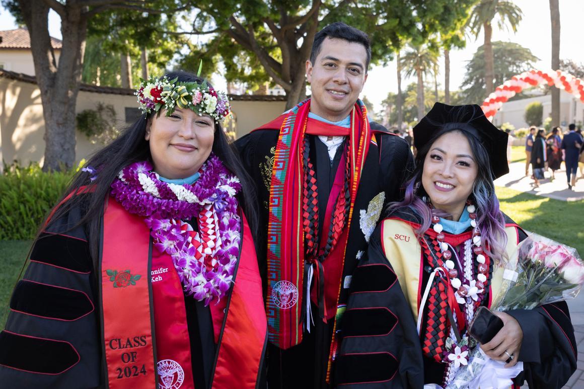 Three graduates in regalia smiling at commencement, with leis, stoles, and flowers.