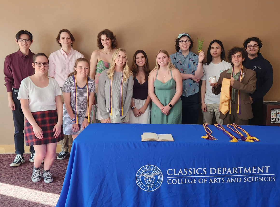 12 students behind a table with a royal blue tablecloth wearing purple and gold honor cords as 2023 Eta Sigma Phi inductees