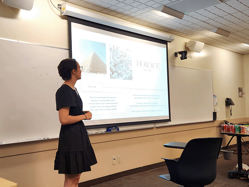 Female student in grey sweater and black skirt giving a presentation in front if a screen