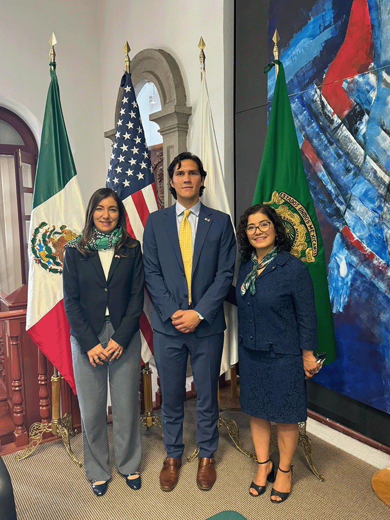 Octavio De Leon standing with two women in front of flags.
