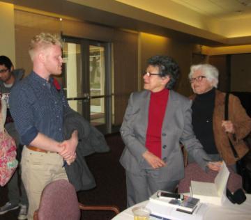 Two individuals are conversing at a table while others look on.