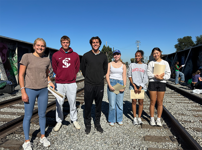 Six students standing near railroad track with blue sky in the background.