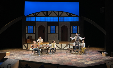 Set for the Book of Will at SCU with 2 men seated and a woman standing with hands on hips at a table on the left and a woman seated and a man standing with arms overhead at a table on the right