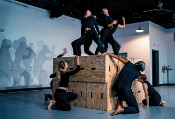 Dance students in black tops and leggings climbing an jumping off 4 large wooden boxes with their shdows projected in a white wall behind