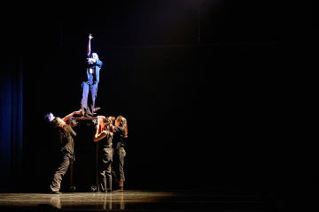 Dancer in black standing on top of an upended box in a spotlight with 3 others reaching up