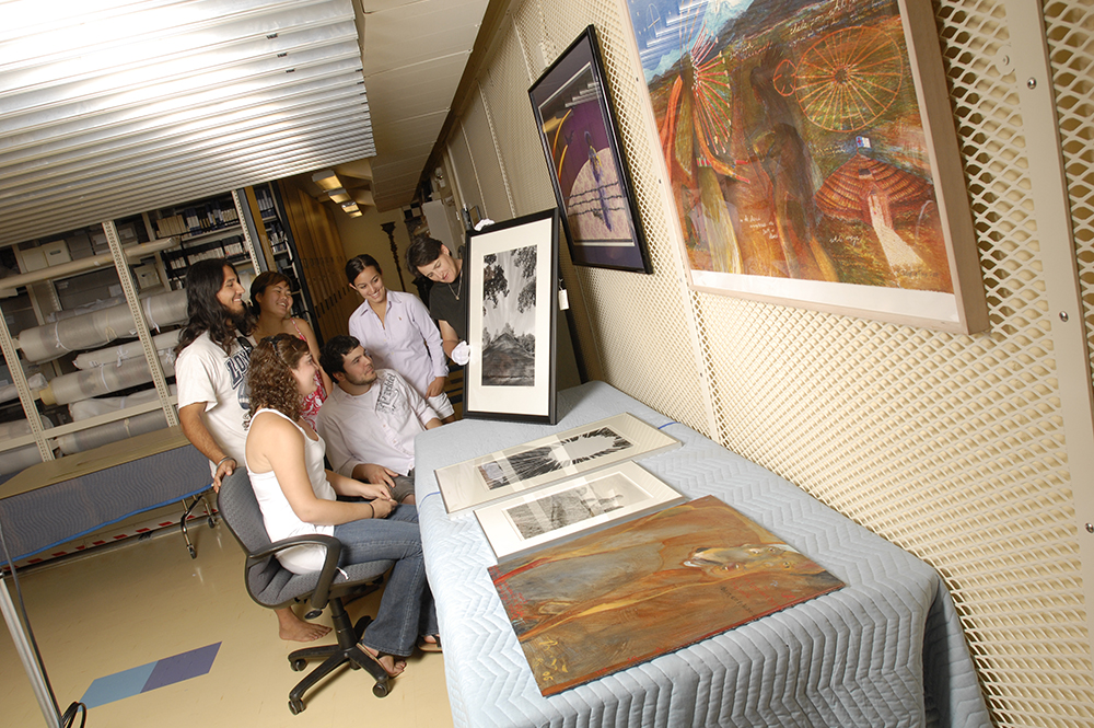 Students gathered around artwork being held by faculty within a collection storage room