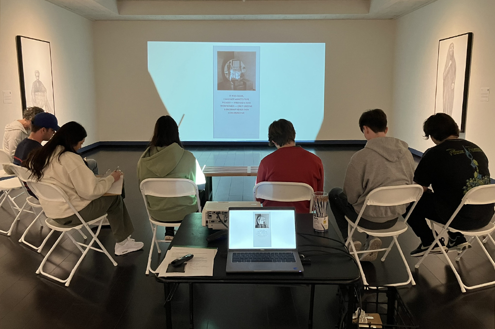 Students seated in gallery with projection of artwork on wall in front of them and physical artwork hanging on the walls to their sides