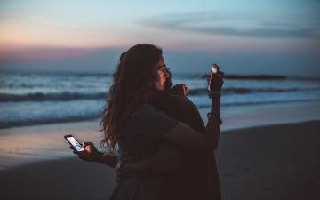 couple embracing on the beach, each looking at mobile device screens