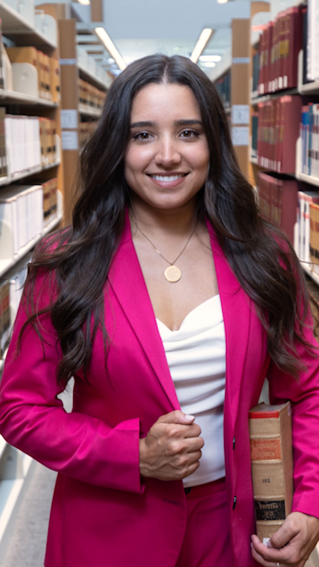 Mirelle Raza, a Santa Clara Graduate, smiling while holding a book and grasping her blazer in a library