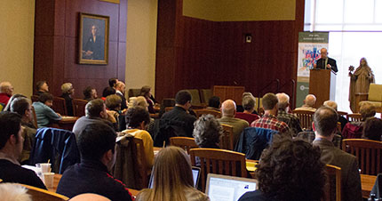 A conference room filled with attendees listening to a speaker at a podium.