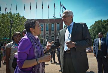 Man in suit speaking to woman holding a microphone outside. Several flags are visible.