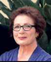 Woman wearing glasses and a blue sweater with green foliage in the background.