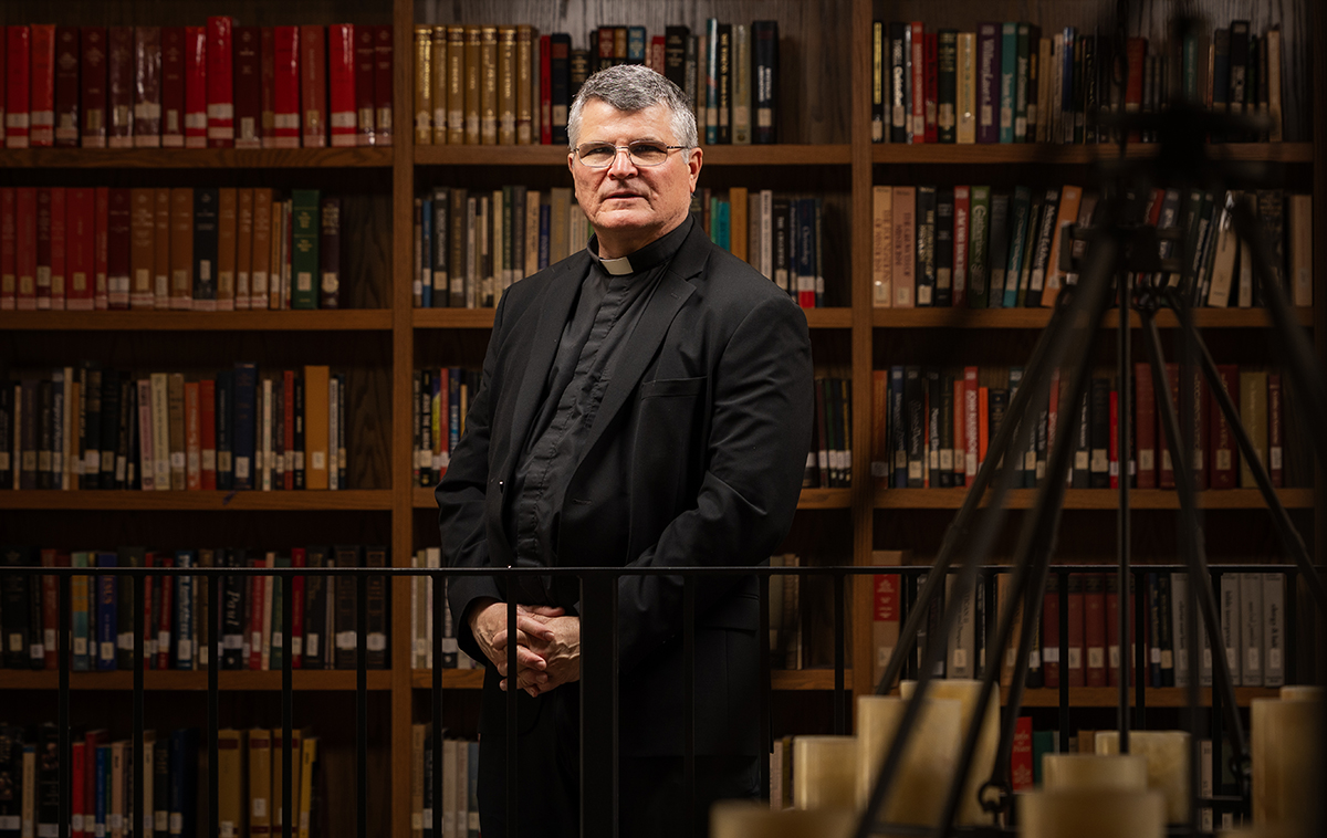 Paul Mariani stands in front of library shelves with chandelier in the foreground.