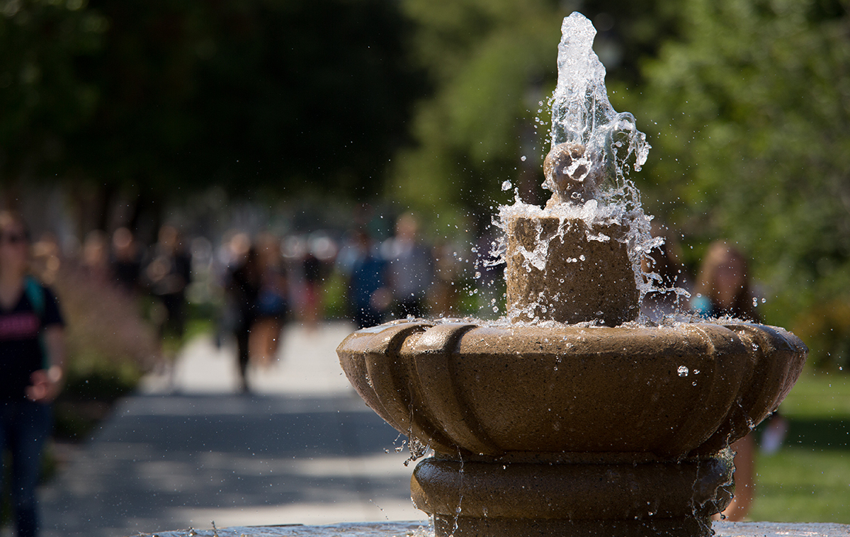 A close-up of a running water fountain with out-of-focus students in the background.