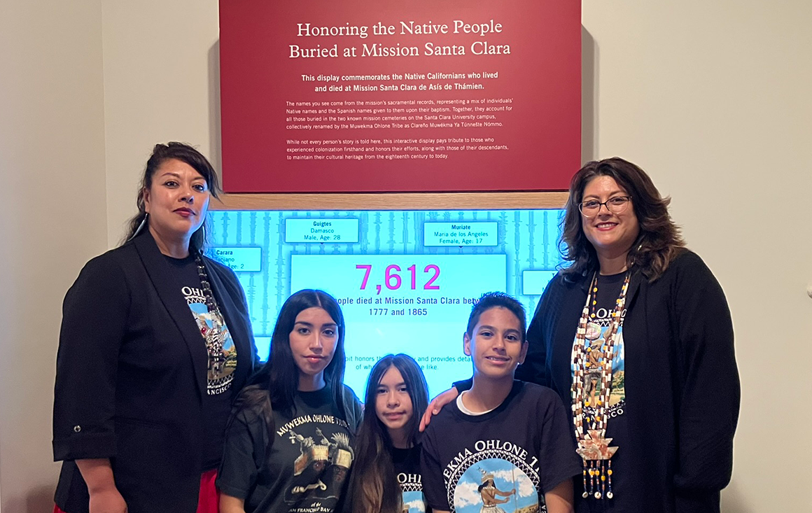 A group of Muwekma Ohlone women and youth stand in front of a digital screen in a museum gallery listing the number of indigenous people buried at Mission Santa Clara.