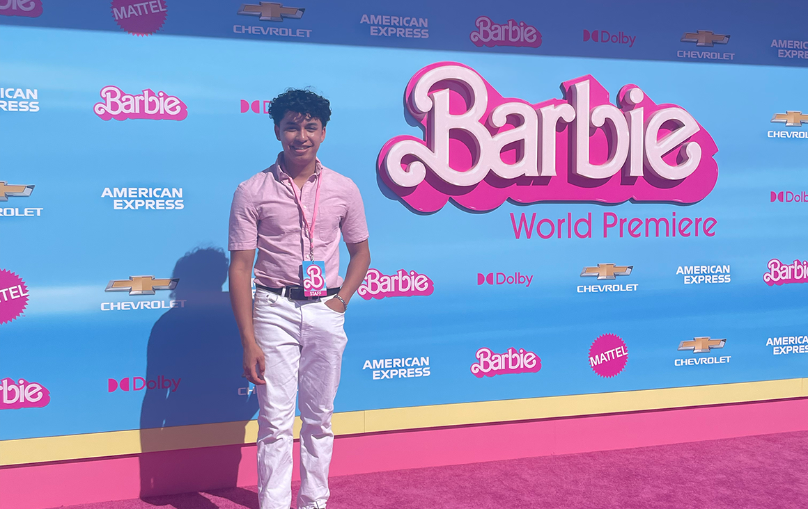 A man in a pink button-up stands on a pink carpet in front of a Barbie promotional film poster.
