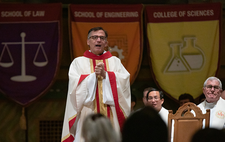 Fr. Kevin O'Brien speaks in the Mission Church with academic flags hanging in the background