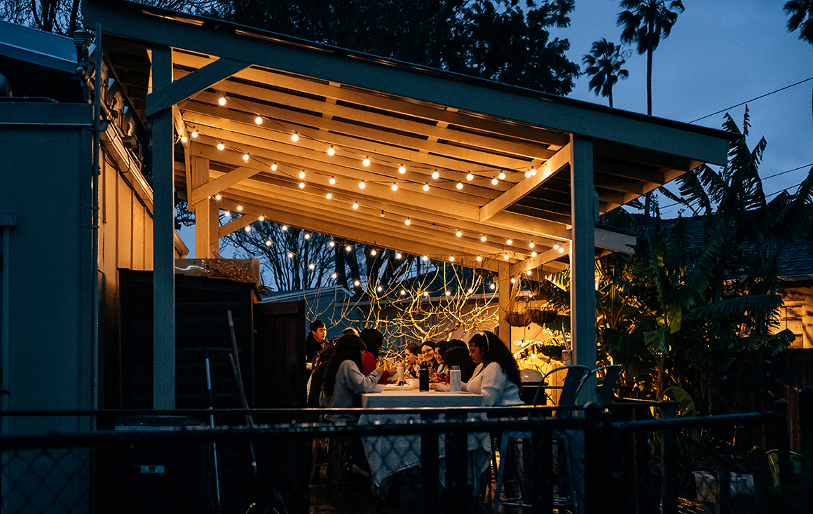 A group of students gather around a dinner table in an outdoor kitchen at night illuminated by warm string lights.