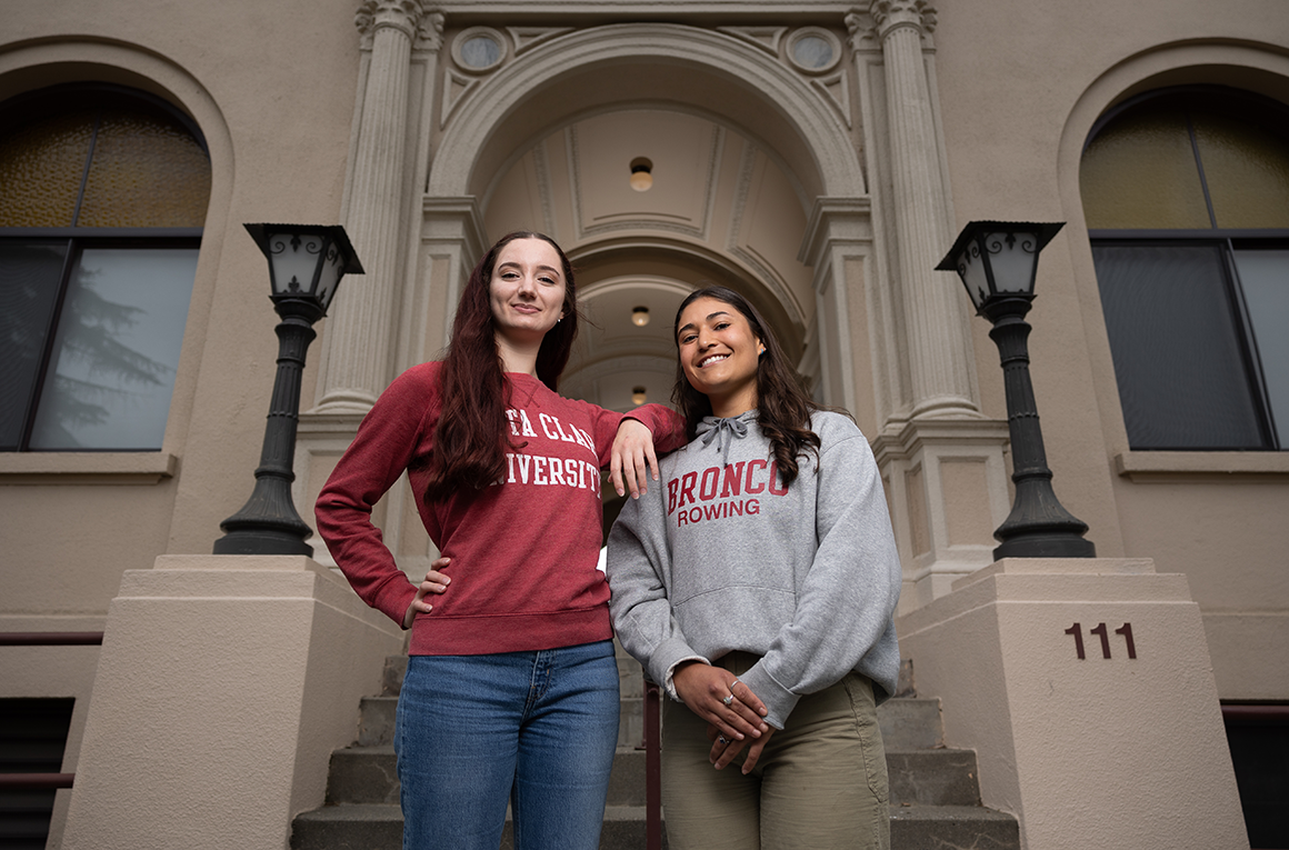 Two students stand at the foot of a staircase by a building's arched entrance. image link to story
