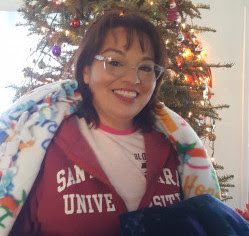 A person smiling, wrapped in blankets, with a Christmas tree in the background.