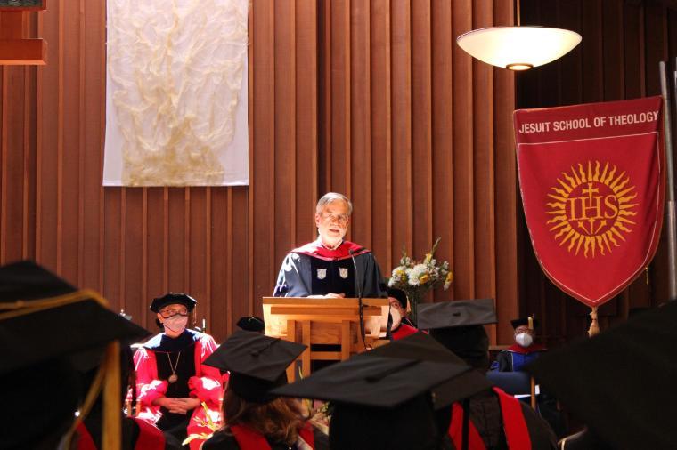 Arthur Holder speaking at a commencement ceremony with audience and banners.