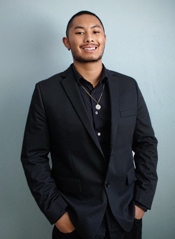 A person wearing a black suit smiling in front of a plain background.