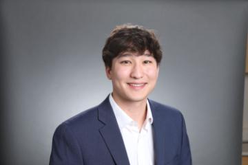 A man in a suit smiling in front of a grey background.