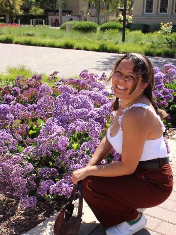 A person smiling, kneeling by purple flowers in a garden.