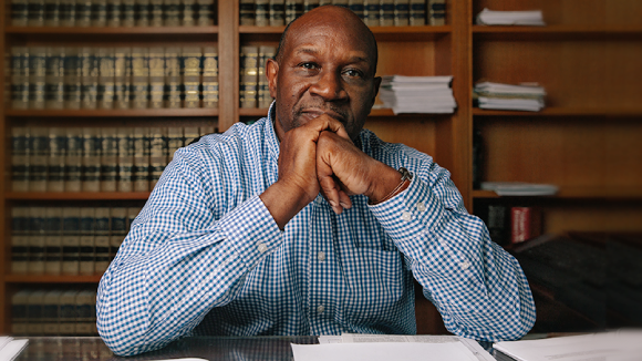 A bald Black man in a blue button-down shirt sits at a desk with his hands steepled under his chin