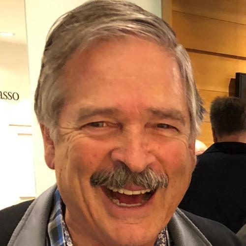 A person smiling warmly while wearing a checkered shirt and jacket indoors.