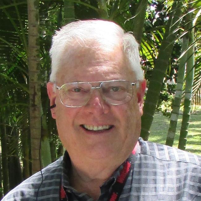 A person smiling outdoors with greenery in the background.