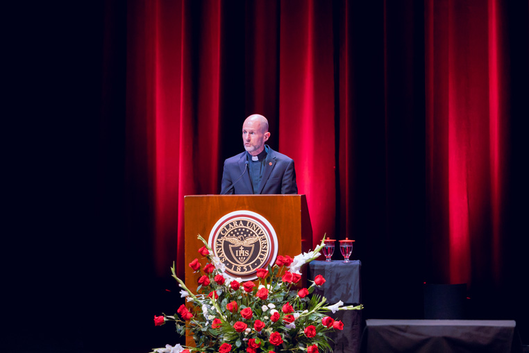 Fr Carnes speaks in a clerical collar at a podium with floral decorations