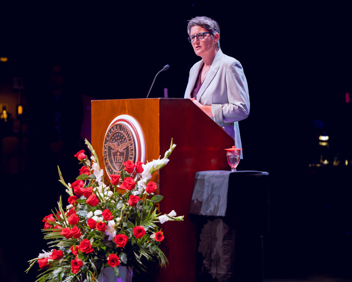 Kate Morris in a light suit at a podium with floral decorations