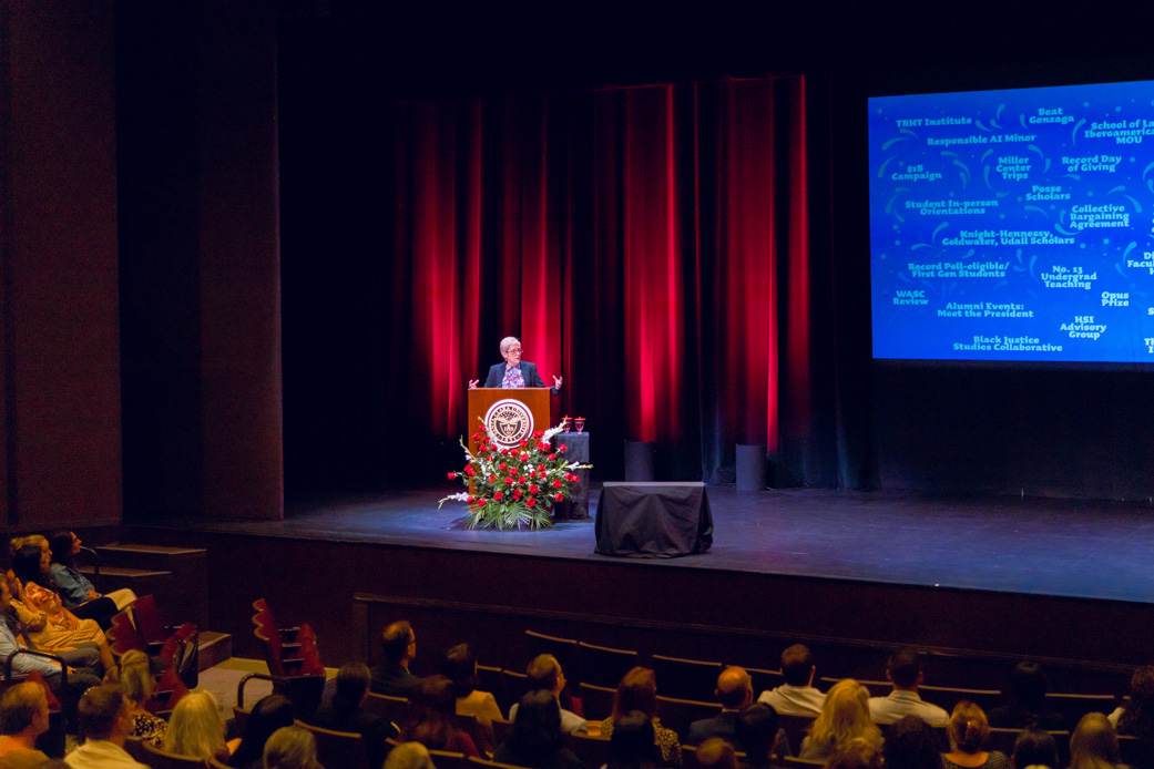 Fr. Carnes at a podium on stage with a projection screen, addressing an audience