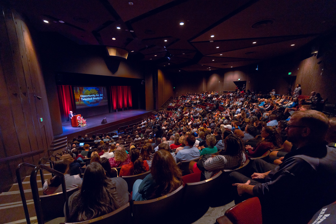 Wide view of President Sullivan on stage with a full audience, screen displays 'Opportunity for All Talented Students.