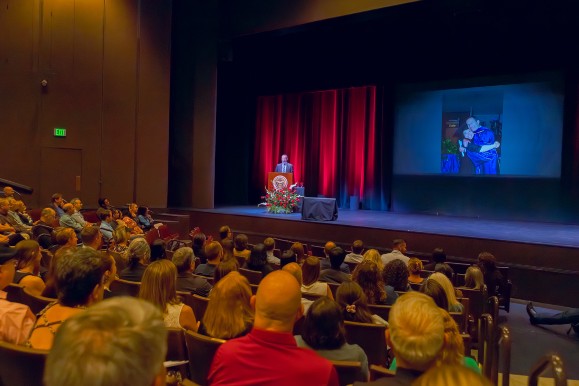 Provost Glaser at a podium, audience watching a projected image of his daughter graduating