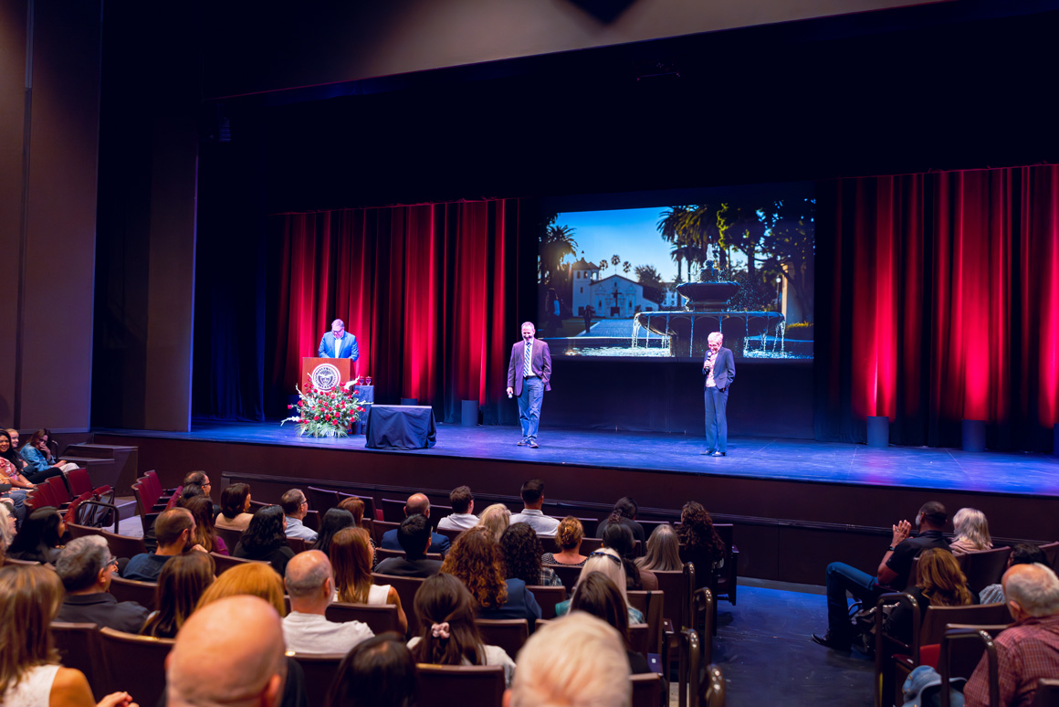 Ed Ryan, Provost Glaser, and President Sullivan on stage, large screen shows a university fountain and mission church