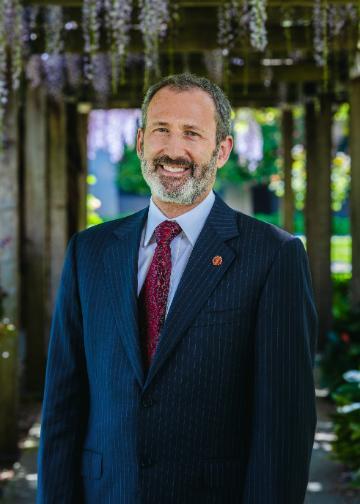 Photo of James Glaser. Man in a navy suit and red tie standing under an arbor.