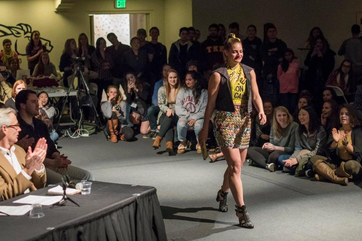 A model wearing a pencil skirt and vest walks down the runway in front of a crowd