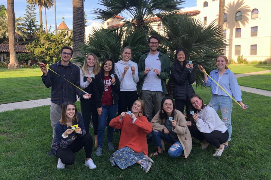 A group of students smiling and posing with tape measures and badges in front of trees on campus