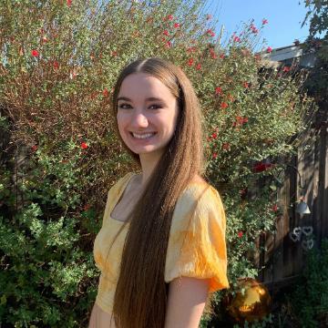 A person in a yellow dress smiling outdoors with plants in the background.