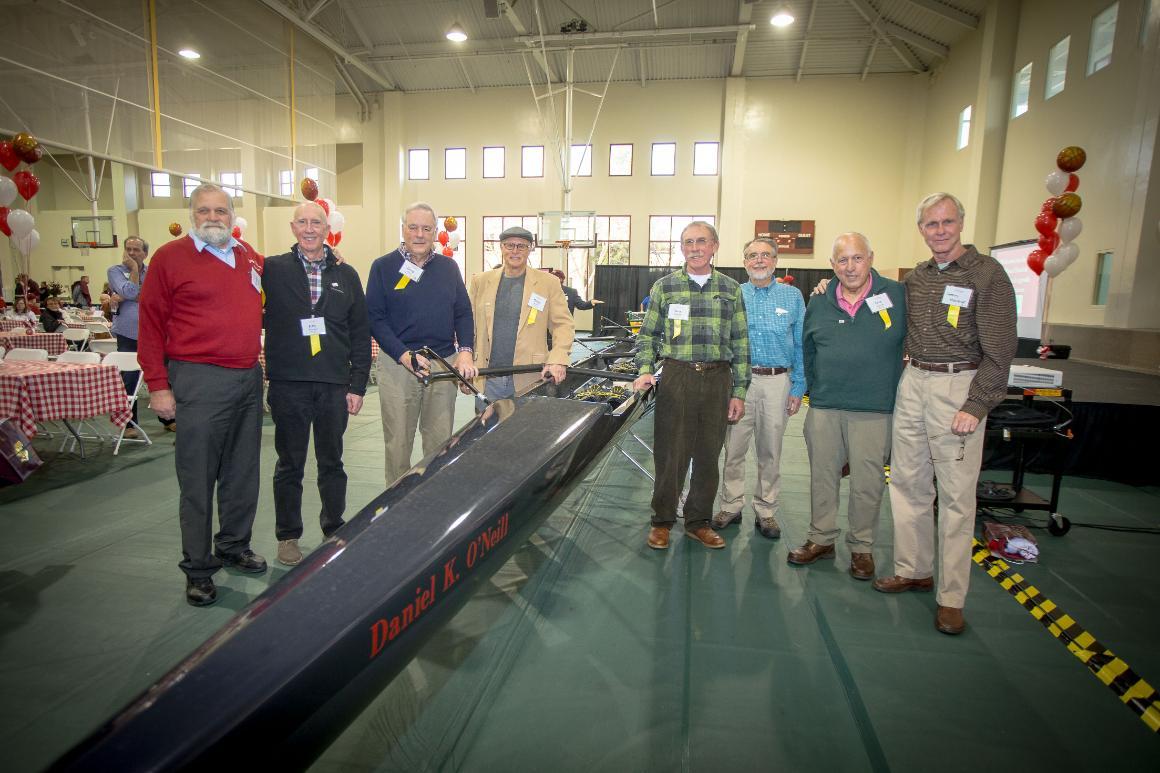 Eight men in their early 70s standing around a rowing shell, smiling.