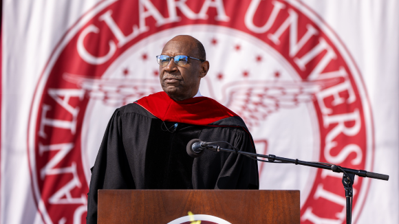 A Black man in glasses and commencement regalia standing at a podium in front of the Santa Clara University seal.